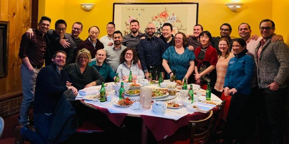 Image of a diverse group of twenty-one adults sitting and standing behind a round table after finishing dinner, posing for a group photo