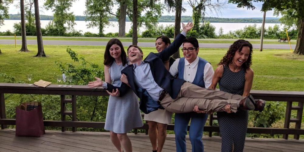 Image of a young man being held up in a horizontal position by four young women who stand behind him, all dressed for a summer cocktail party. Everyone in the photo is laughing.